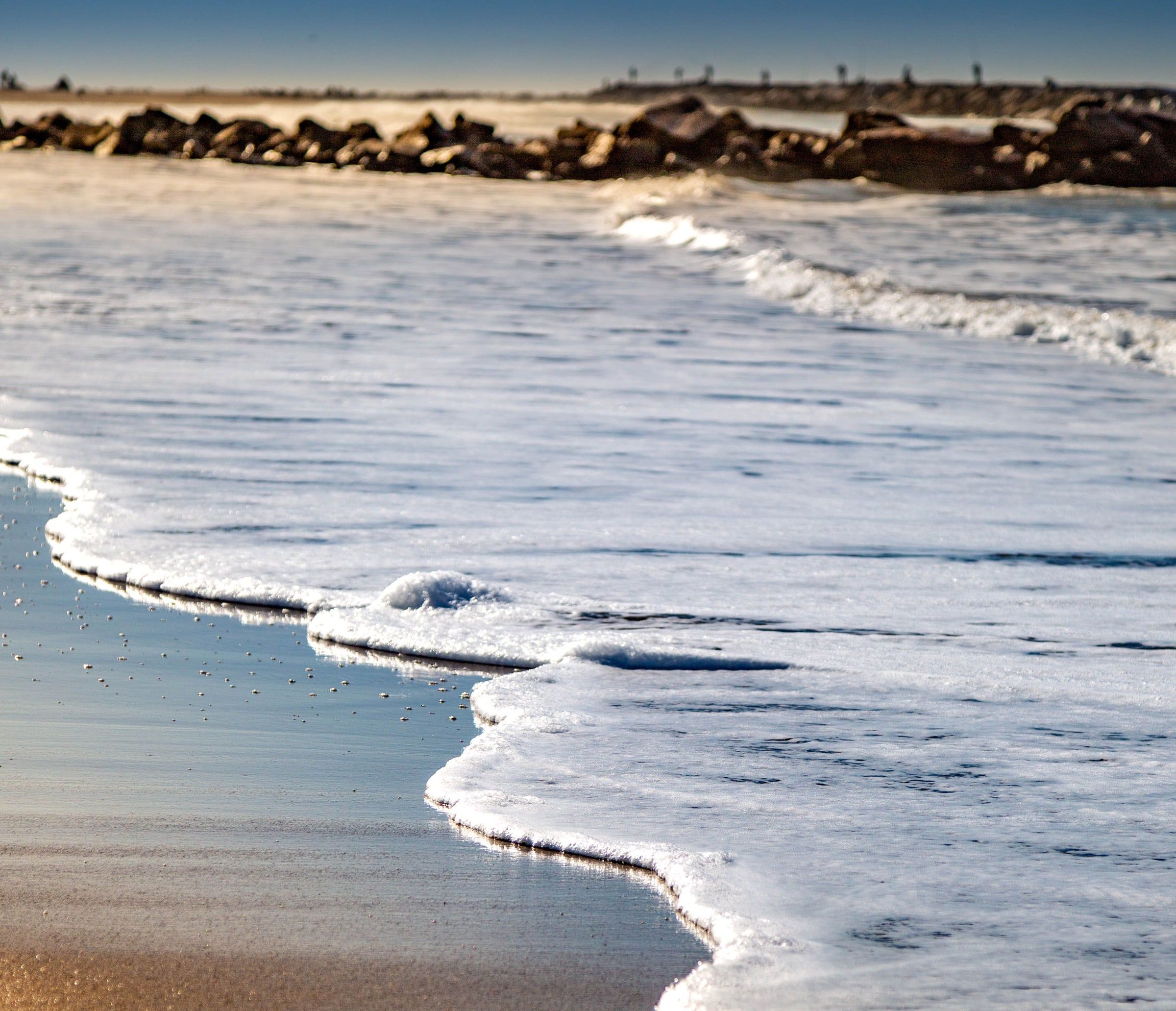A wave washing up on the sand, with a rock jetty behind
